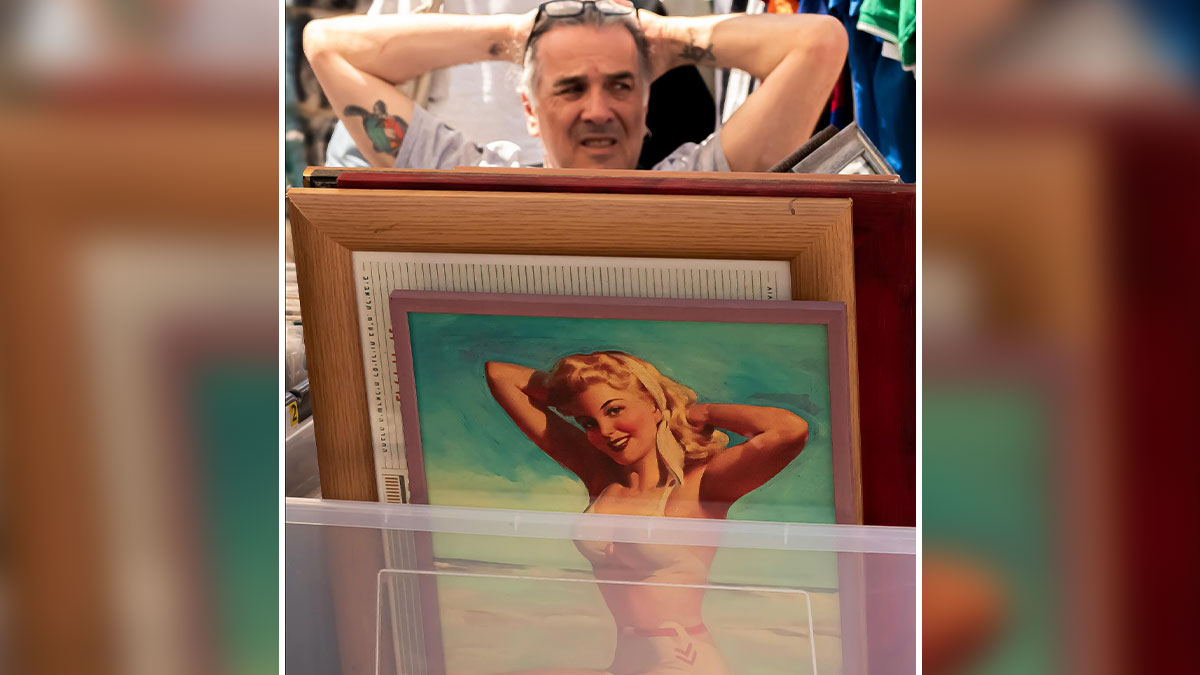Man with tattoo relaxing behind vintage framed photos at an outdoor market in a powerful street photo of everyday life