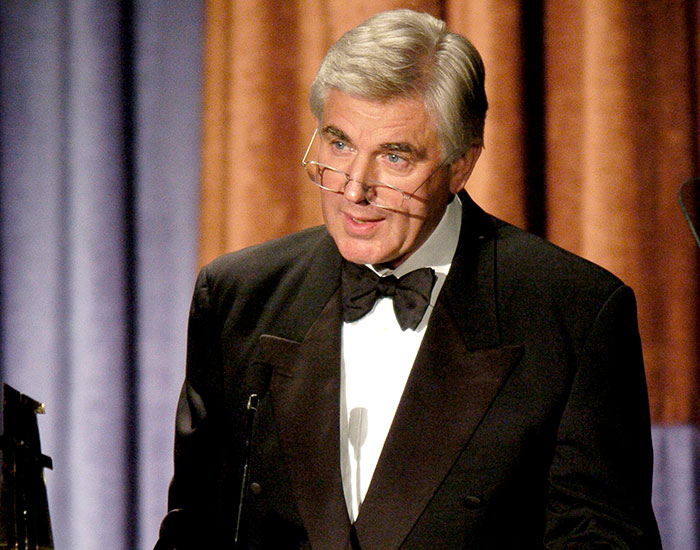 Man in a tuxedo wearing glasses, speaking at a formal event, symbolizing J.K. Rowling team addressing viral cancer rumor. Man in a tuxedo wearing glasses, speaking at a formal event, symbolizing J.K. Rowling team addressing viral cancer rumor.