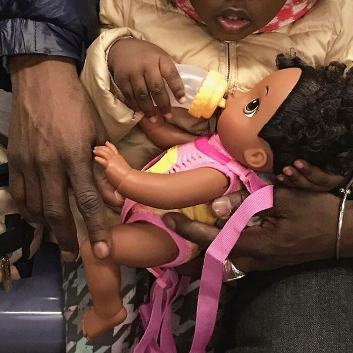 Child on New York subway holding a doll wearing a pink outfit, being fed with a baby bottle.