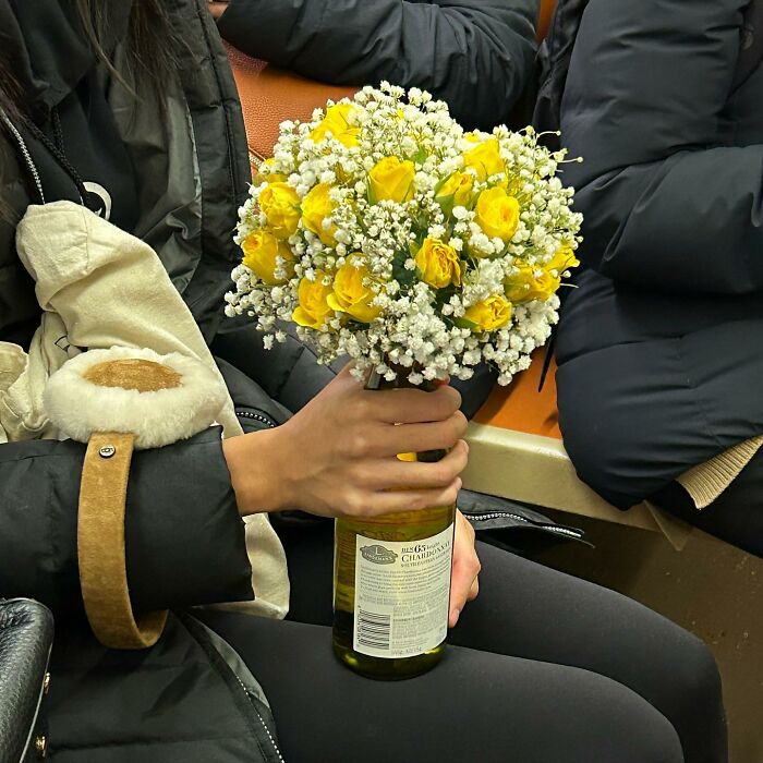Person on New York subway holding a wine bottle with a bouquet of yellow and white flowers inside.