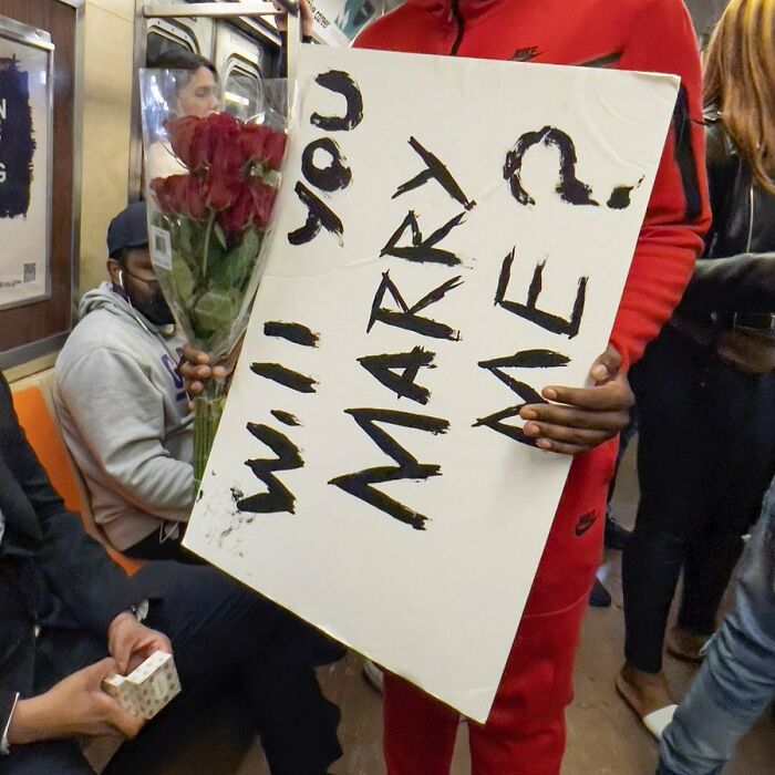 Person on New York subway holding a sign that says Will You Marry Me with a bouquet of roses.