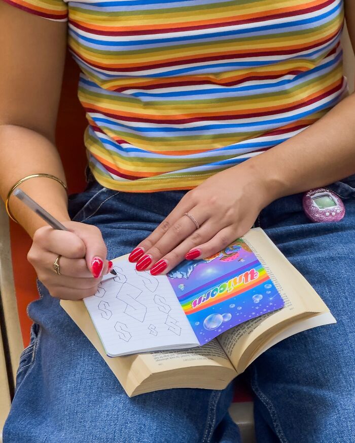 Person in striped shirt and jeans drawing in notebook while sitting with book and a digital pet on subway in New York.