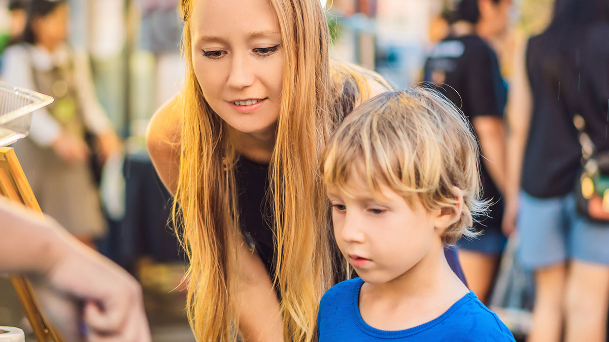 Kid with nut allergy looking at a sample while a worker offers it to him at a busy outdoor market scene.