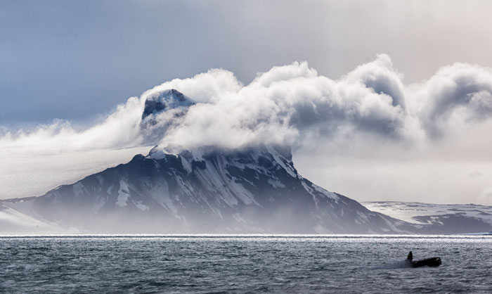 Mountain partially covered by clouds over cold ocean waters with a small boat, illustrating the 1831 mystery of the sun turning blue. Mountain partially covered by clouds over cold ocean waters with a small boat, illustrating the 1831 mystery of the sun turning blue.