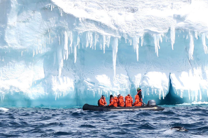 Researchers in orange jackets on a boat near large icy cliffs under a bright blue sky exploring the 1831 mystery of the sun turning blue. Researchers in orange jackets on a boat near large icy cliffs under a bright blue sky exploring the 1831 mystery of the sun turning blue.