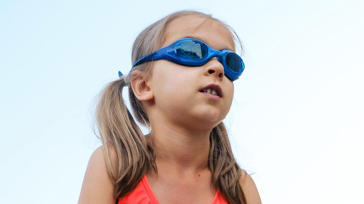 Young girl wearing goggles and modest swimwear preparing for a swim lesson with male swim teacher present nearby.