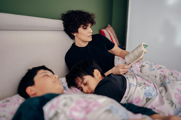 Woman reading a book in bed while two children sleep, illustrating hotel guests disturbing neighbors with late-night ruckus.