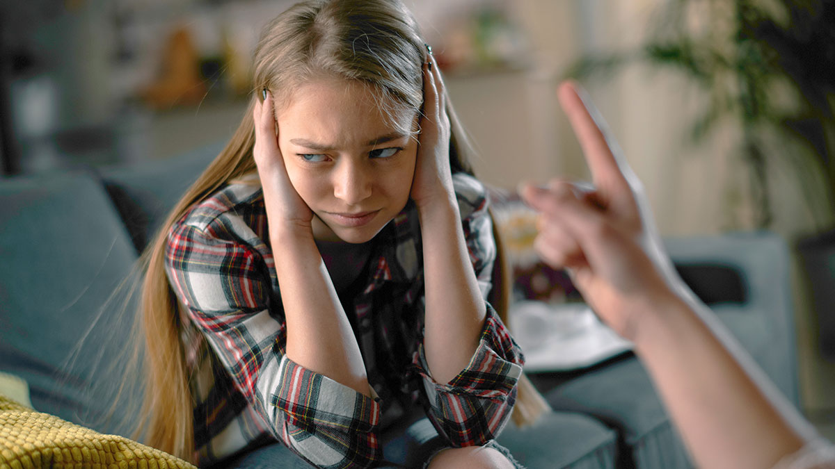 Teen girl covering ears looking upset while adult points finger, representing dad pulling funding for trip conflict.