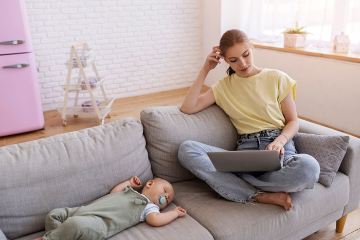 Teen girl using laptop on couch while baby sibling lies nearby, reflecting on dad pulling funding for trip after party choice. Teen girl using laptop on couch while baby sibling lies nearby, reflecting on dad pulling funding for trip after party choice.