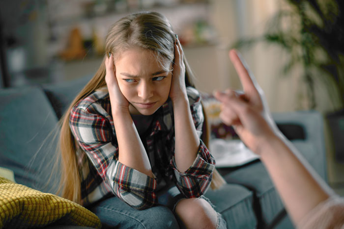 Teen girl covering ears while sitting on couch during a tense conversation about dad pulling funding for her trip. Teen girl covering ears while sitting on couch during a tense conversation about dad pulling funding for her trip.