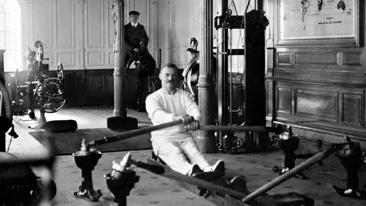 Man rowing on vintage gym rowing machine in early 1900s setting, rare historical photo of Titanic era interior.