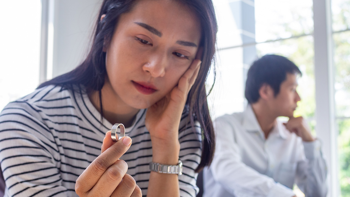 Woman holding a wedding ring looking sad while a man sits in the background, representing cheating and divorce issues.