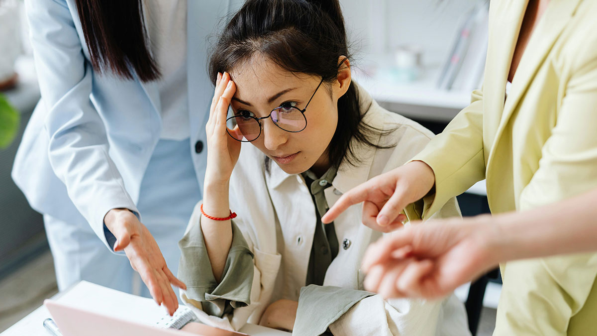 Stressed employee covering her face while coworkers angrily point fingers in a tense toxic work environment.