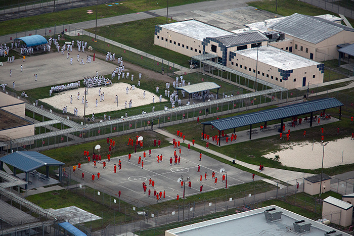 Aerial view of detainees in an ICE detention center yard with fences and multiple recreation areas visible. Aerial view of detainees in an ICE detention center yard with fences and multiple recreation areas visible.