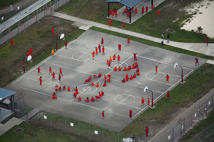 Aerial view of detainees in orange uniforms gathered on an outdoor basketball court at an ICE detention facility. Aerial view of detainees in orange uniforms gathered on an outdoor basketball court at an ICE detention facility.