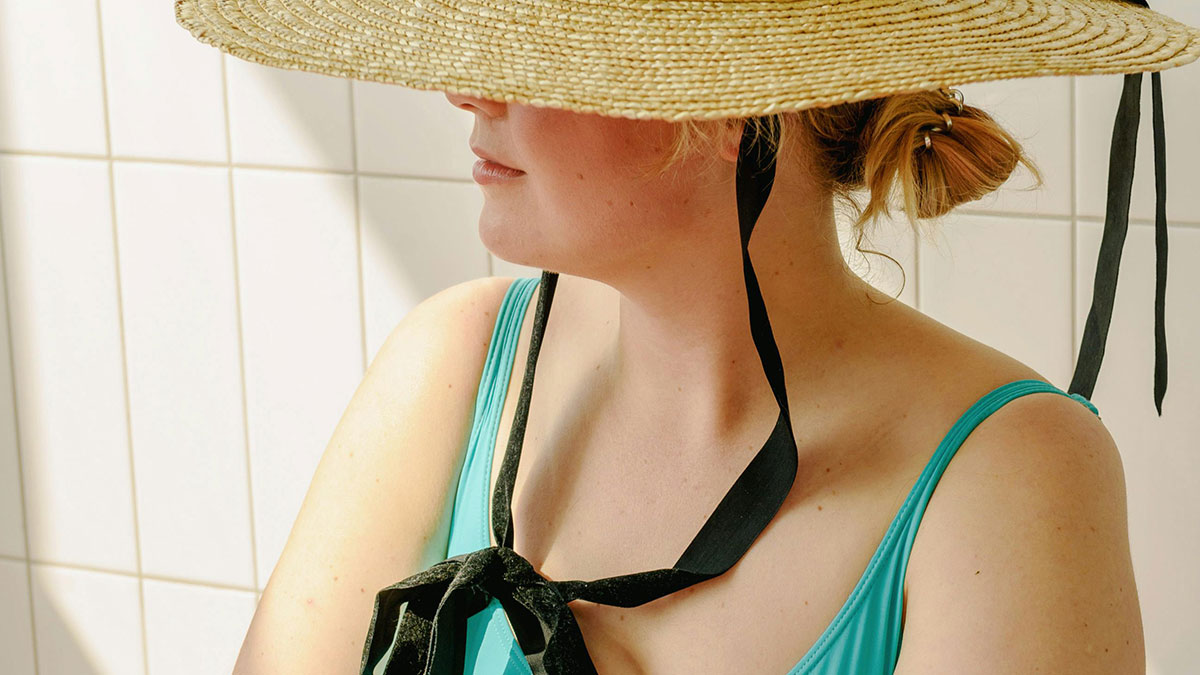 Woman in turquoise swimsuit and wide straw hat resting indoors, reflecting on vacations that ended up being trips to hell.