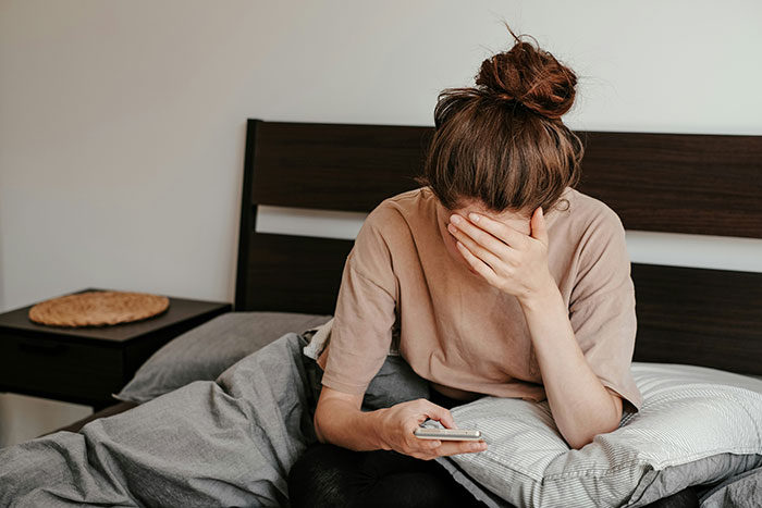 Bride-to-be sitting on bed upset, covering face with hand while holding phone, reacting to future MIL branding her stupid. Bride-to-be sitting on bed upset, covering face with hand while holding phone, reacting to future MIL branding her stupid.