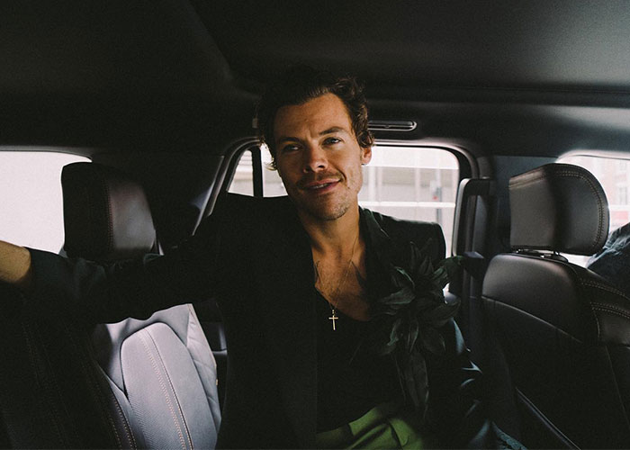 Harry Styles sitting inside a car wearing a black jacket and a cross necklace, casually smiling at the camera. Harry Styles sitting inside a car wearing a black jacket and a cross necklace, casually smiling at the camera.