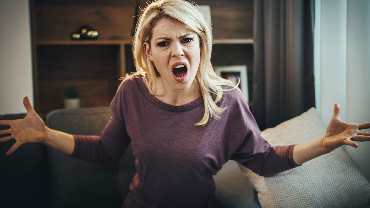 Angry wife with blonde hair gesturing in frustration on a couch, expressing emotions related to husband and ashes honeymoon incident.
