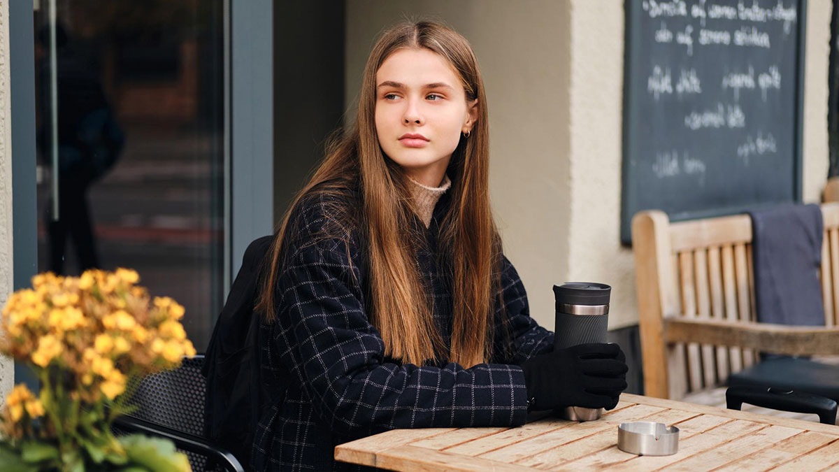 Young woman with allergies sitting outside cafu00e9 holding a cup, facing rigid cafu00e9 rules during coffee date.