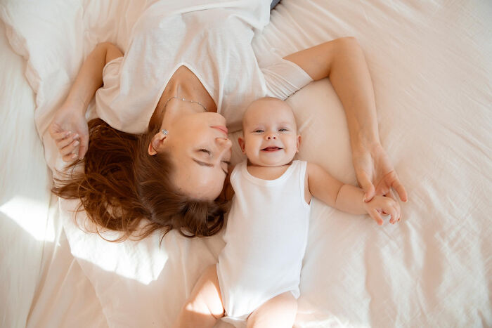 Mother lying on bed holding hands with smiling baby, highlighting toxic man relationship issues from two women perspective.