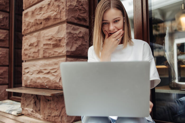 Young woman shocked while reading about toxic man romantically involved with two women on laptop outdoors.