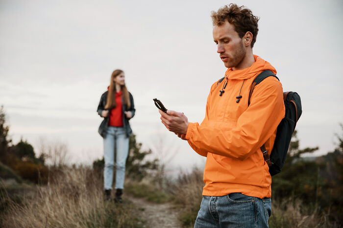 Young man in an orange jacket texting on phone outdoors while a woman stands in the background, illustrating toxic man behavior.