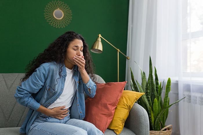 Young woman sitting on sofa, holding stomach and covering mouth, showing signs of illness and suspicious behavior concerns. Young woman sitting on sofa, holding stomach and covering mouth, showing signs of illness and suspicious behavior concerns.
