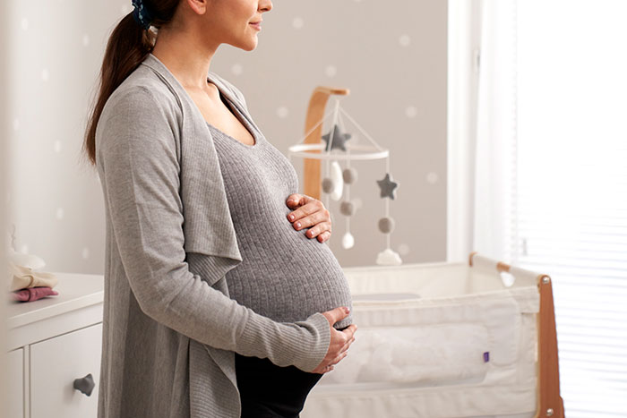 Pregnant woman in gray sweater holding belly in softly lit nursery, symbolizing a woman's world crumbling after a dark secret.
