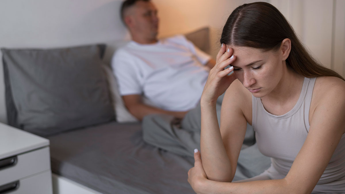 A woman looking distressed and contemplative, sitting on a bed while her husband sits silently in the background.