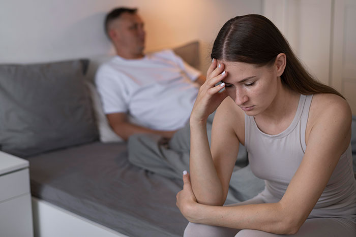 A woman sitting on the edge of a bed looking distressed while her husband sits in the background upset after a dark secret revelation.