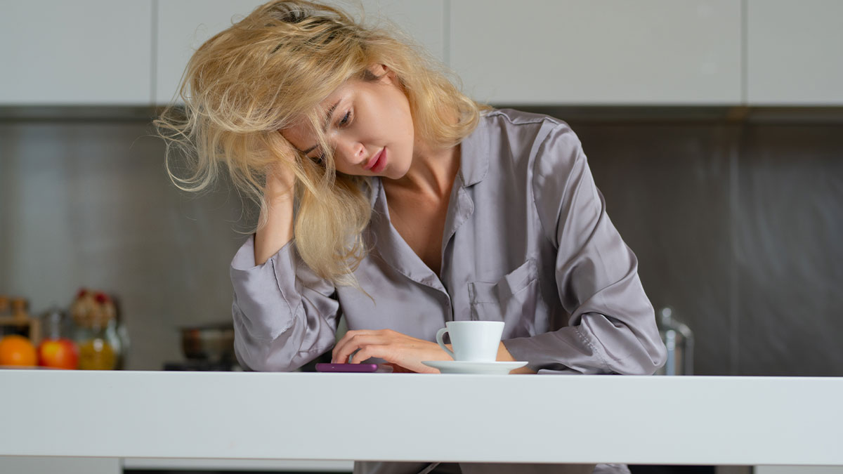 Tired woman in pajamas looking distressed at her phone in kitchen, reflecting on challenges of motherhood and fears.