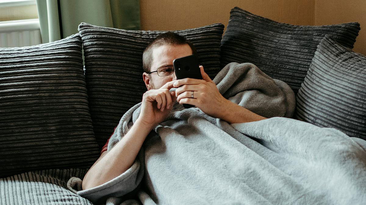 Man wrapped in a blanket on a couch, using a smartphone, reflecting themes of a toxic marriage and family abandonment.