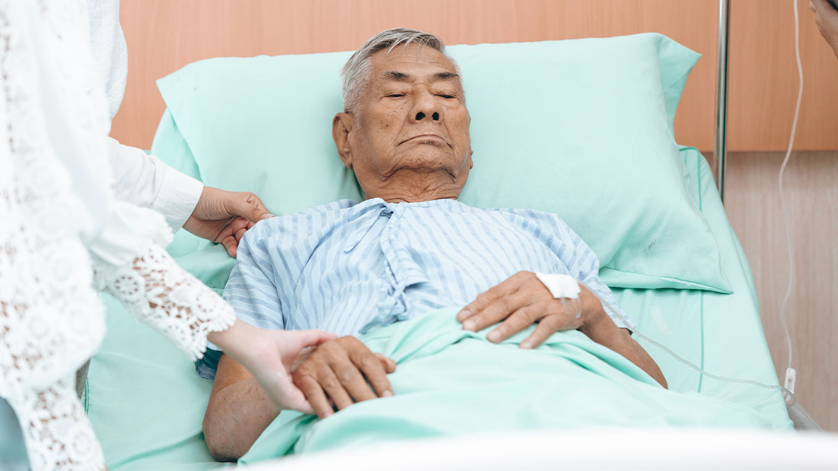 Older man lying in hospital bed with visitor holding his hand showing compassion and care.