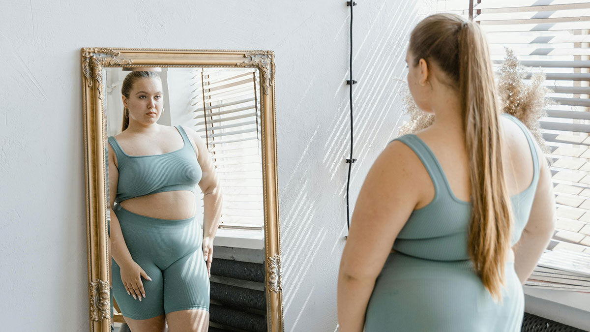 Woman in activewear looking at her reflection in a mirror, reflecting themes of new weight and emotional struggle.