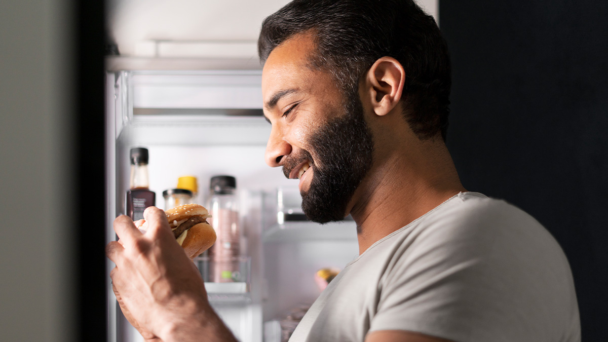 Man smiling while holding a burger in front of an open refrigerator, illustrating living with men habits.