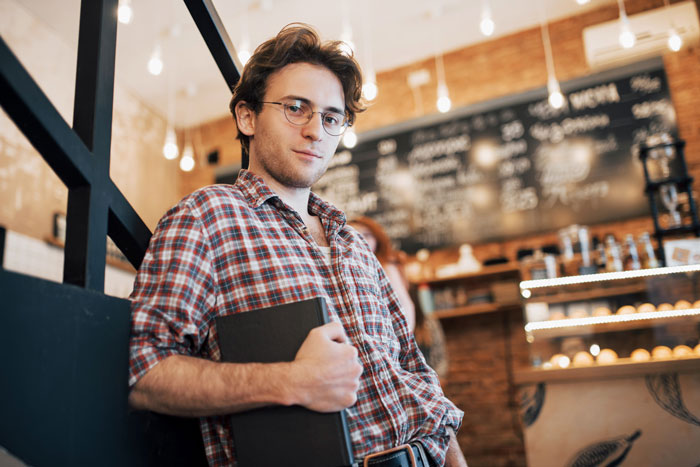 Teen holding a book, leaning against a wall in a café, reflecting on babysitting half siblings and secret plans. Teen holding a book, leaning against a wall in a café, reflecting on babysitting half siblings and secret plans.