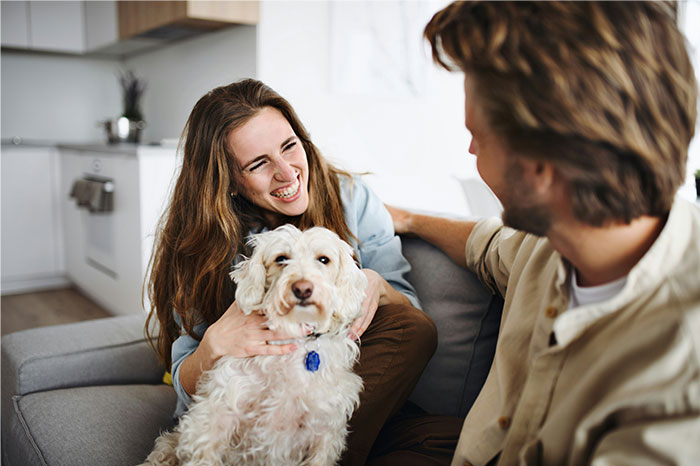 Two friends laughing and sitting on a couch with a dog, illustrating ways people got permanently banned from a friend's house.
