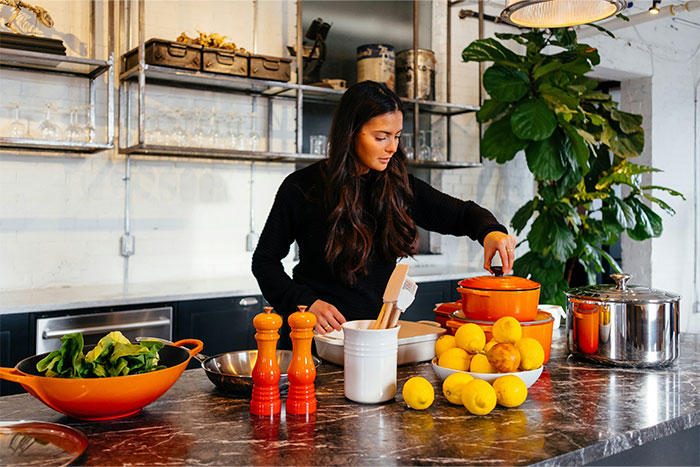 Woman preparing food in modern kitchen with lemons and orange cookware, illustrating ways people got banned from friend’s house.
