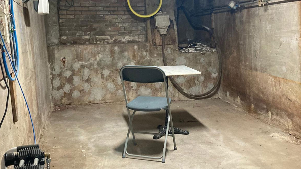 Dilapidated break room with concrete walls, a single chair and table, resembling a prison environment.