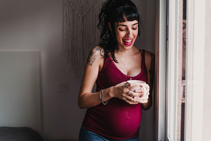 Pregnant woman smiling by window, holding a cup, reflecting on keeping baby and male best friend stepping in father role. Pregnant woman smiling by window, holding a cup, reflecting on keeping baby and male best friend stepping in father role.