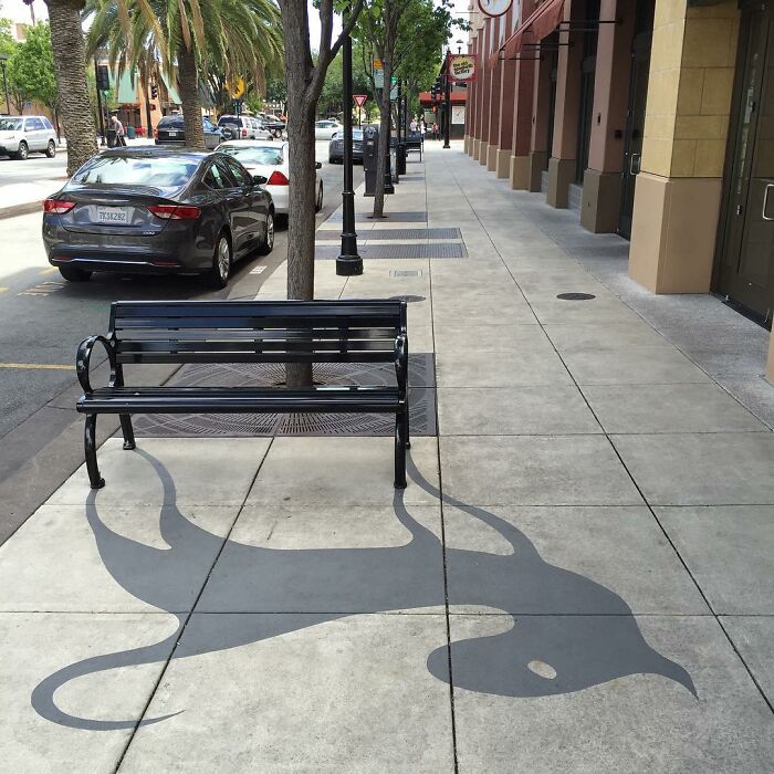 Shadow art installation on sidewalk creatively using bench legs to form a large, abstract creature silhouette at night.