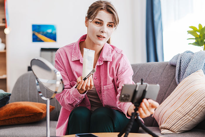Gym influencer recording skincare video at home, showing product and makeup brush while filming on smartphone tripod. Gym influencer recording skincare video at home, showing product and makeup brush while filming on smartphone tripod.