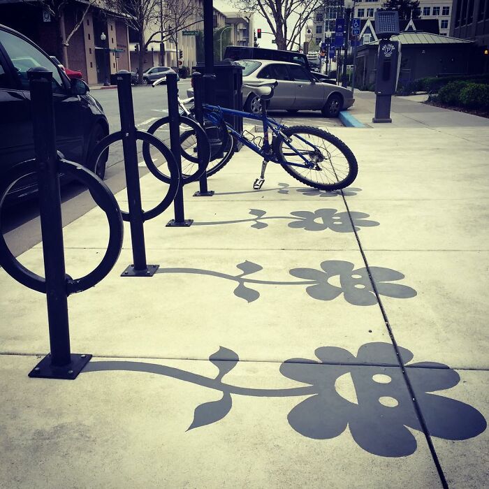 Black metal bike racks casting clever shadow art of smiling flowers on a concrete sidewalk in an urban setting.
