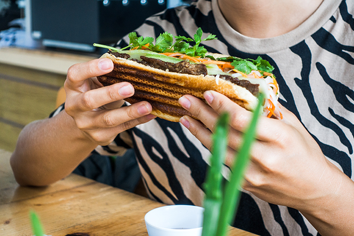 Person holding and about to eat a spicy chicken sandwich topped with fresh herbs and vegetables at a wooden table Person holding and about to eat a spicy chicken sandwich topped with fresh herbs and vegetables at a wooden table
