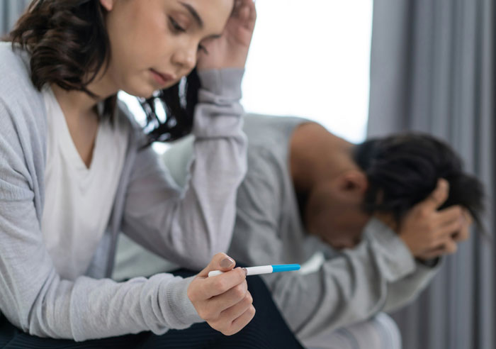 A distressed woman holding a pregnancy test while a man sits in the background with his head in his hands, showing family drama. A distressed woman holding a pregnancy test while a man sits in the background with his head in his hands, showing family drama.