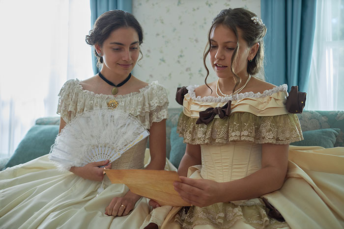 Two women in extravagant ball gowns discussing a letter, highlighting the theme of aesthetic and exclusion.