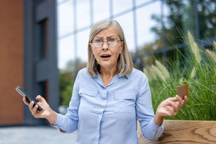 Surprised middle-aged woman holding phone and card outdoors, reacting to parents lied about food allergies to daughter.