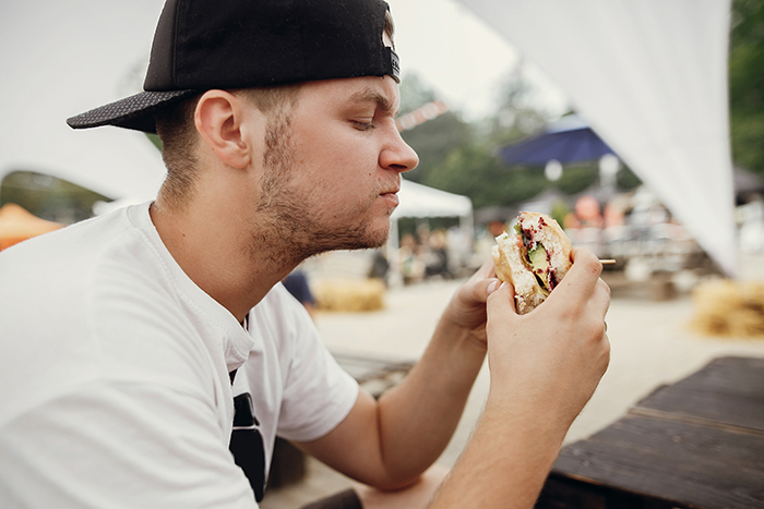 Young man wearing a cap tasting a spicy chicken sandwich outdoors at a casual picnic area on a cloudy day. Young man wearing a cap tasting a spicy chicken sandwich outdoors at a casual picnic area on a cloudy day.