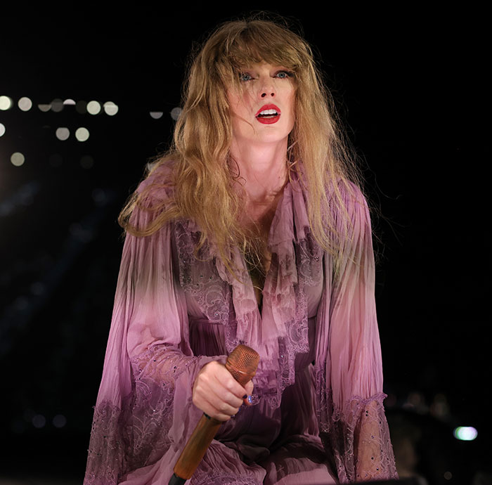 Taylor Swift performing on stage wearing a pink lace dress, holding a microphone under bright concert lights. Taylor Swift performing on stage wearing a pink lace dress, holding a microphone under bright concert lights.